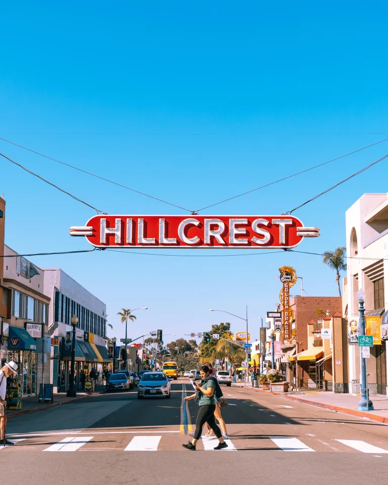 A red sign reading "Hillcrest" hangs above a street lined with shops and people crossing a crosswalk under a clear blue sky.