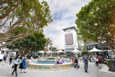 People walk and sit around a tiled fountain in an outdoor UTC Westfield plaza with trees, umbrellas, and a clock tower in the background.