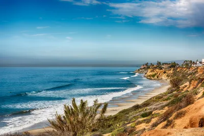 View of a sandy Carlsbad beach with waves rolling in, bordered by rocky cliffs and coastal vegetation under a partly cloudy sky.