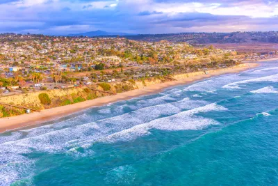 Aerial view of waves hitting Cardiff-by-the-Sea's sandy beach with a coastal town and hills in the background under a partly cloudy sky.
