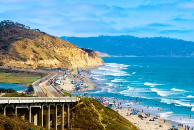 Crowded Torrey Pines State Beach along a coastline with cliffs, a road, and a railway; people are swimming, sunbathing, and walking near the waves under a partly cloudy sky.
