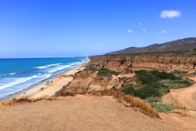 Coastal cliffs overlook the sandy San Onofre beach with scattered people and umbrellas; waves approach shore under a clear blue sky with distant hills in the background.