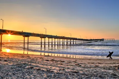 Ocean Beach pier extends over the ocean at sunset, with a surfer carrying a board on the sandy beach in the foreground.