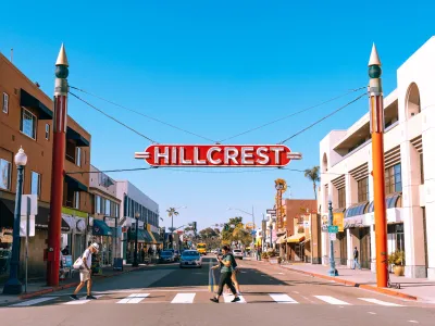 A red sign reading "Hillcrest" hangs above a street lined with shops and people crossing a crosswalk under a clear blue sky.